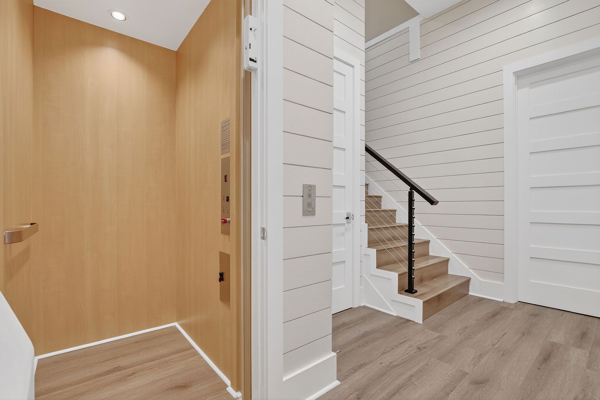 Hallway with wood elevator, stairs with black railing, and white paneled walls.