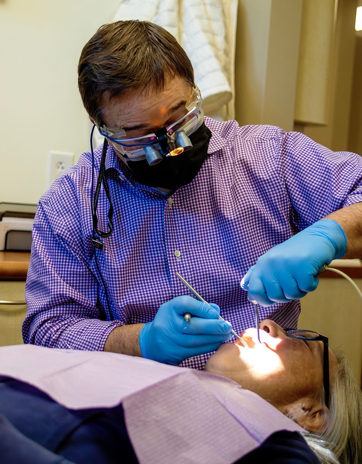 A dentist in a purple patterned shirt, mask, and loupes performs a dental exam on a patient wearing glasses.