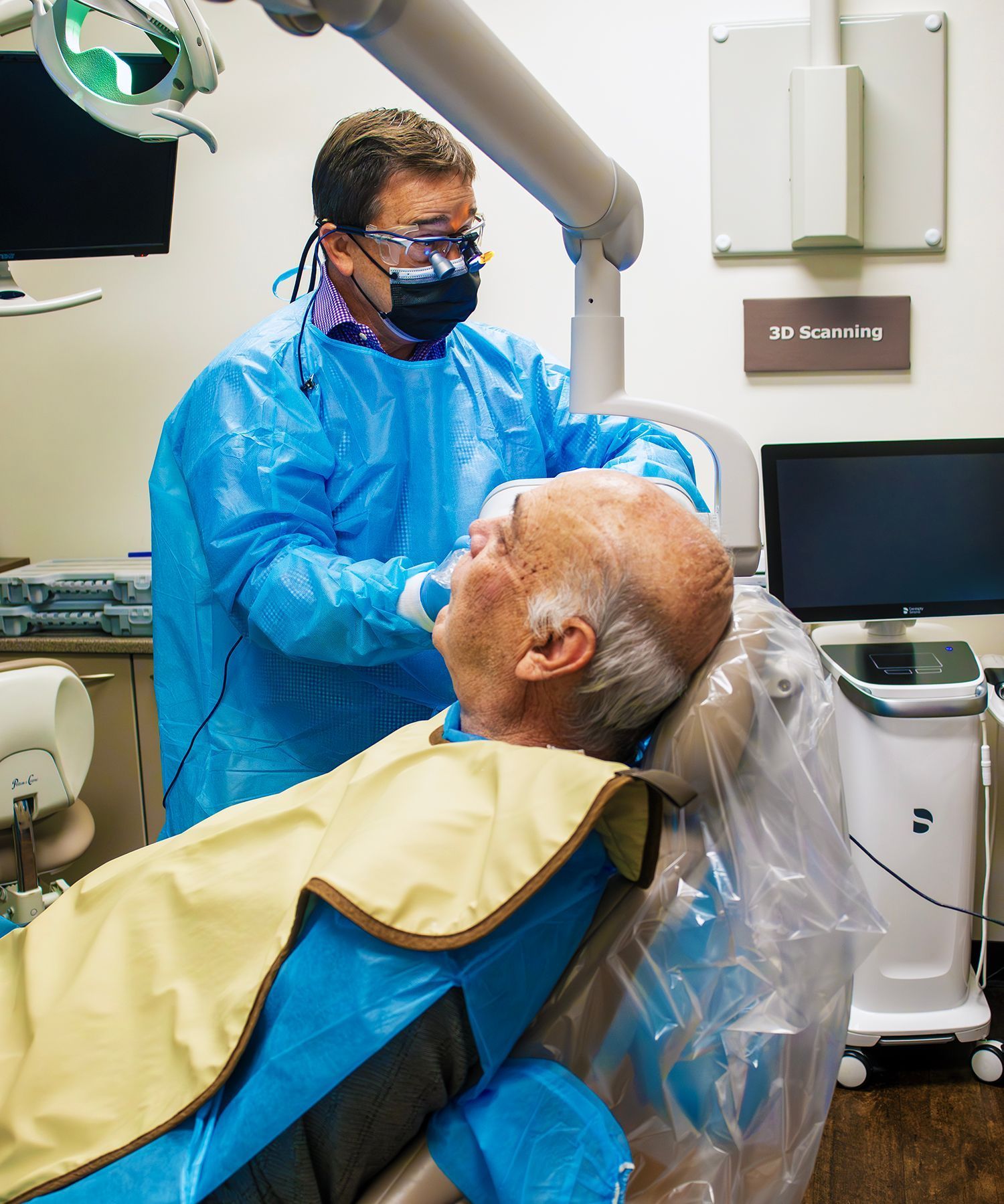 A dentist in protective gear works on a patient lying in a dental chair in a clinical office.