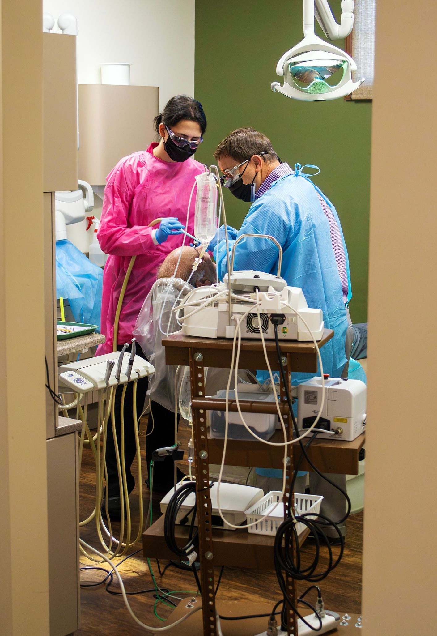 Two medical professionals in scrubs and masks performing a dental procedure on a patient in a clinical office setting.
