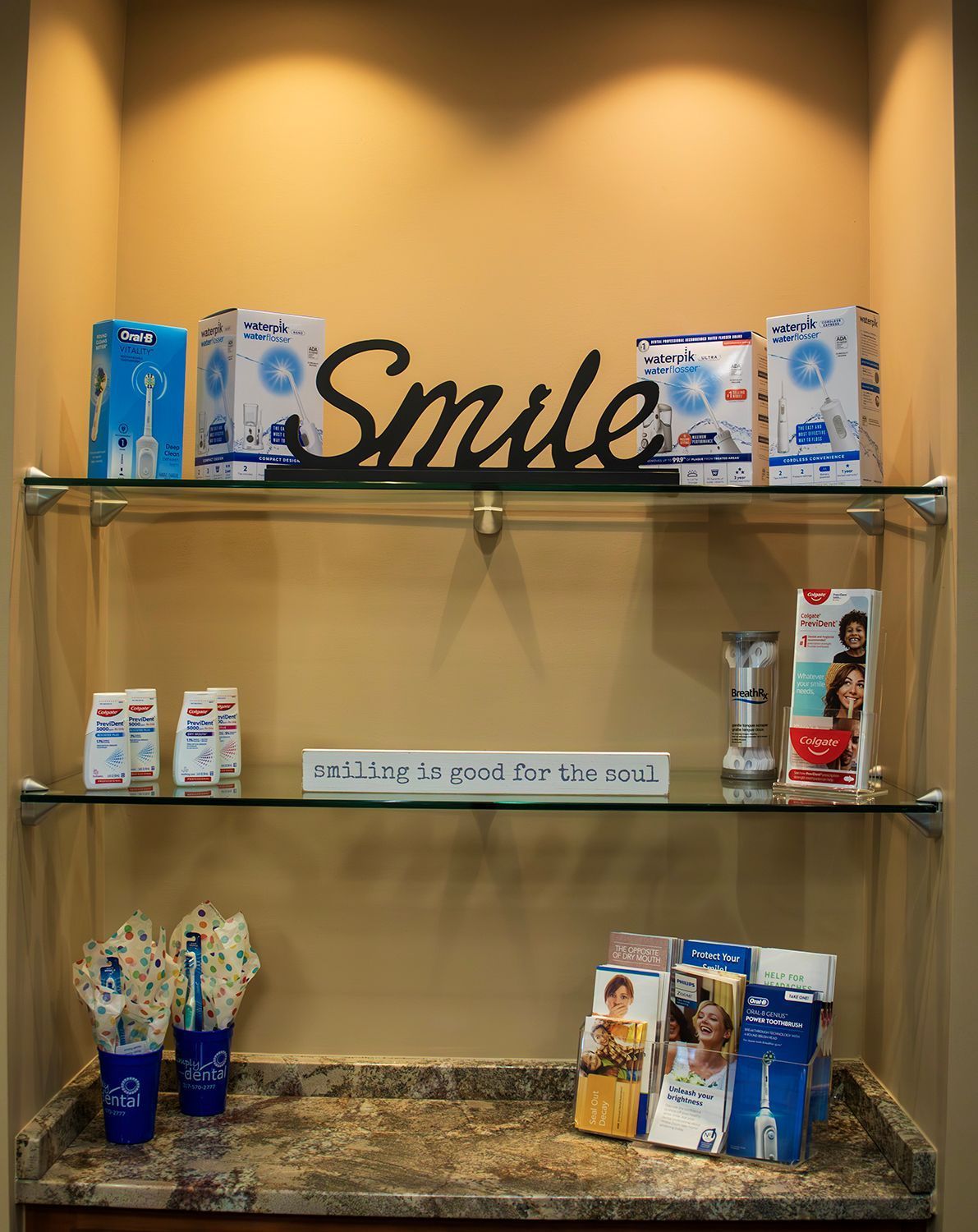 A dental office display shelf features boxes of oral care products, a Smile sign, and pamphlets on a stone countertop.