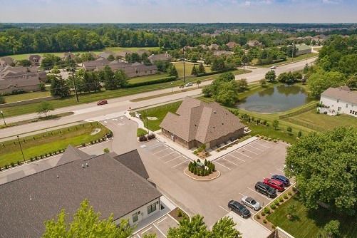 Aerial view of a brown-roofed building, parking lot, road, and pond with surrounding houses and trees.