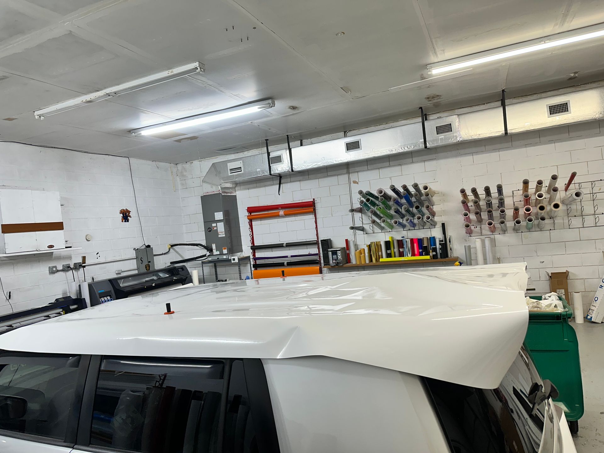 White car in a garage with a large panel resting on its roof, tools and shelves on the wall.