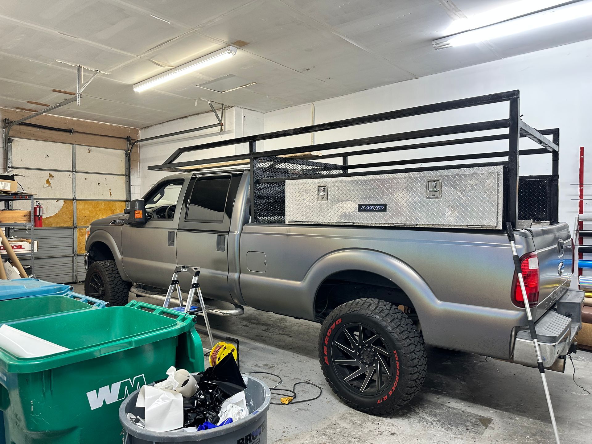 Silver pickup truck in a garage with a black roof rack and side storage boxes, surrounded by tools and bins