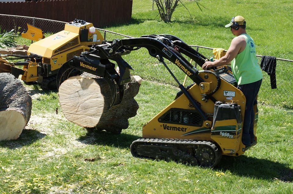 Man operating a yellow mini excavator lifting a large boulder in a grassy yard