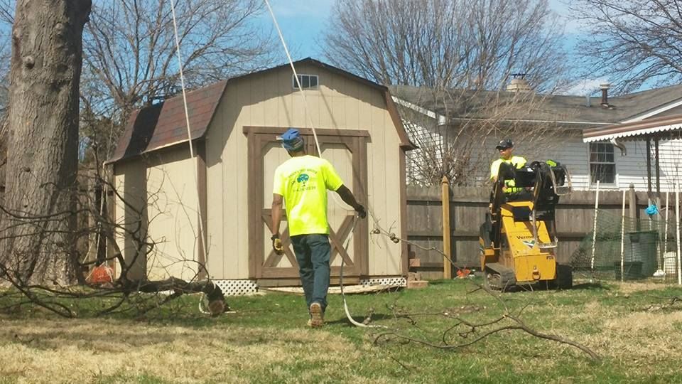 Worker in neon shirt using a hose near a shed while another stands by a wood chipper in a yard