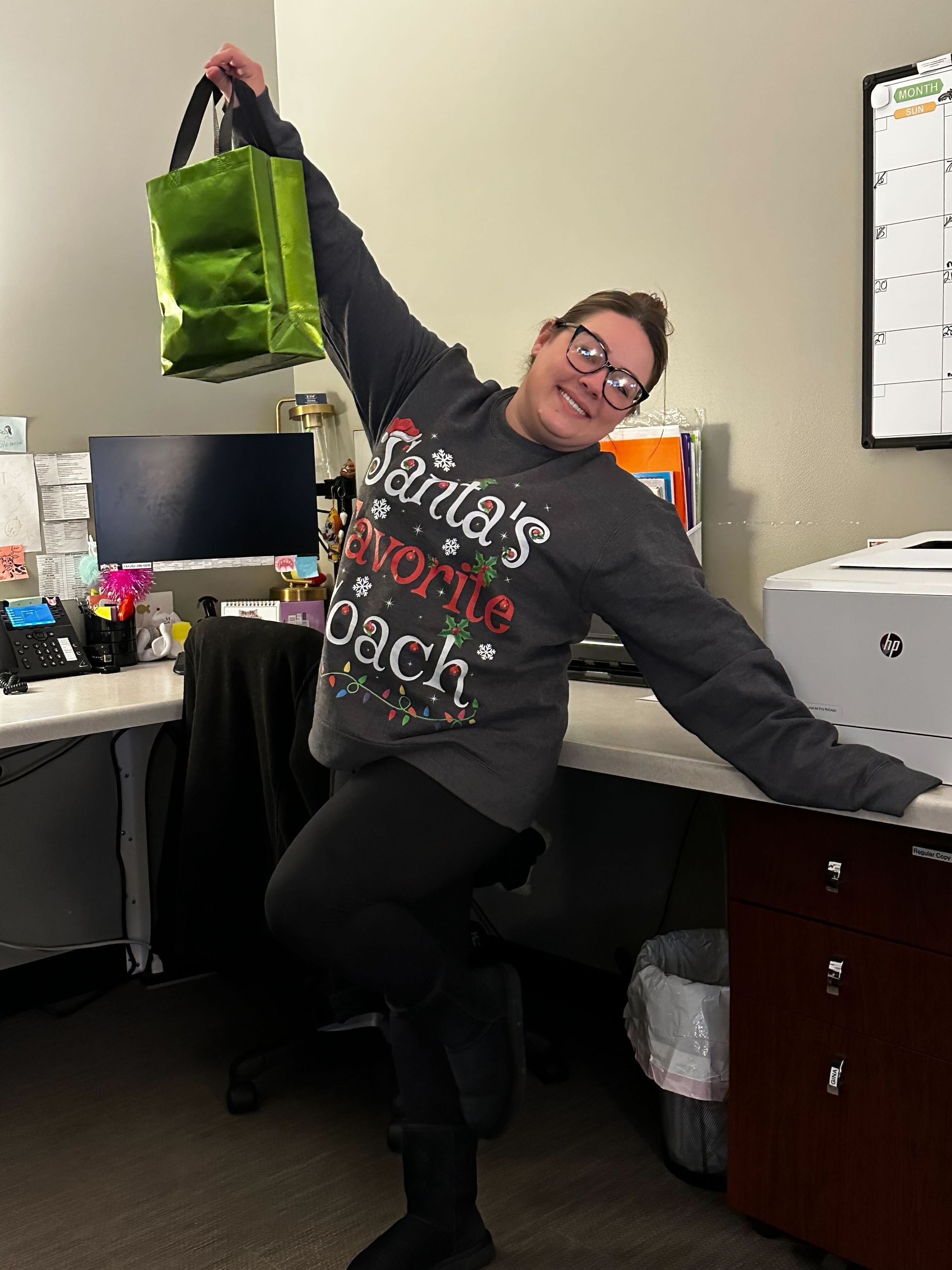 A woman in a santa 's favorite teacher sweater is holding a green bag.