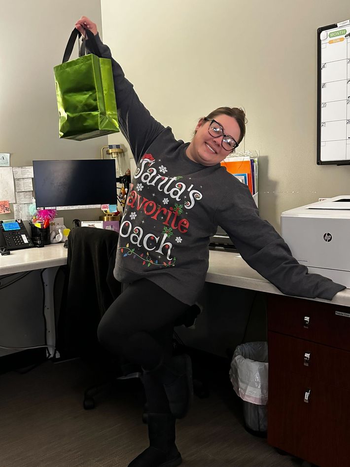 A woman in a santa 's favorite teacher sweater is holding a green bag.