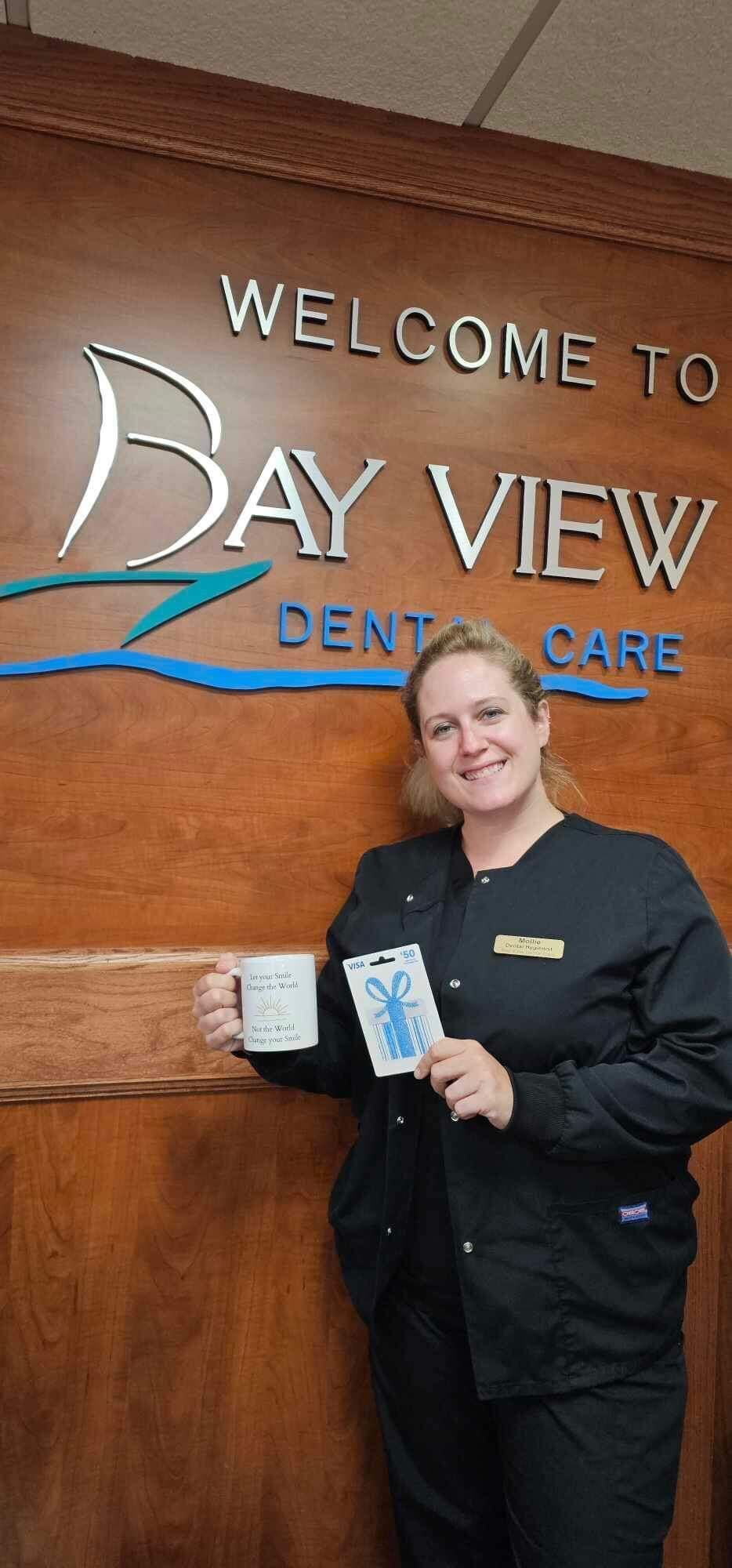 A woman is holding a cup of coffee in front of a sign that says welcome to bay view dental care.