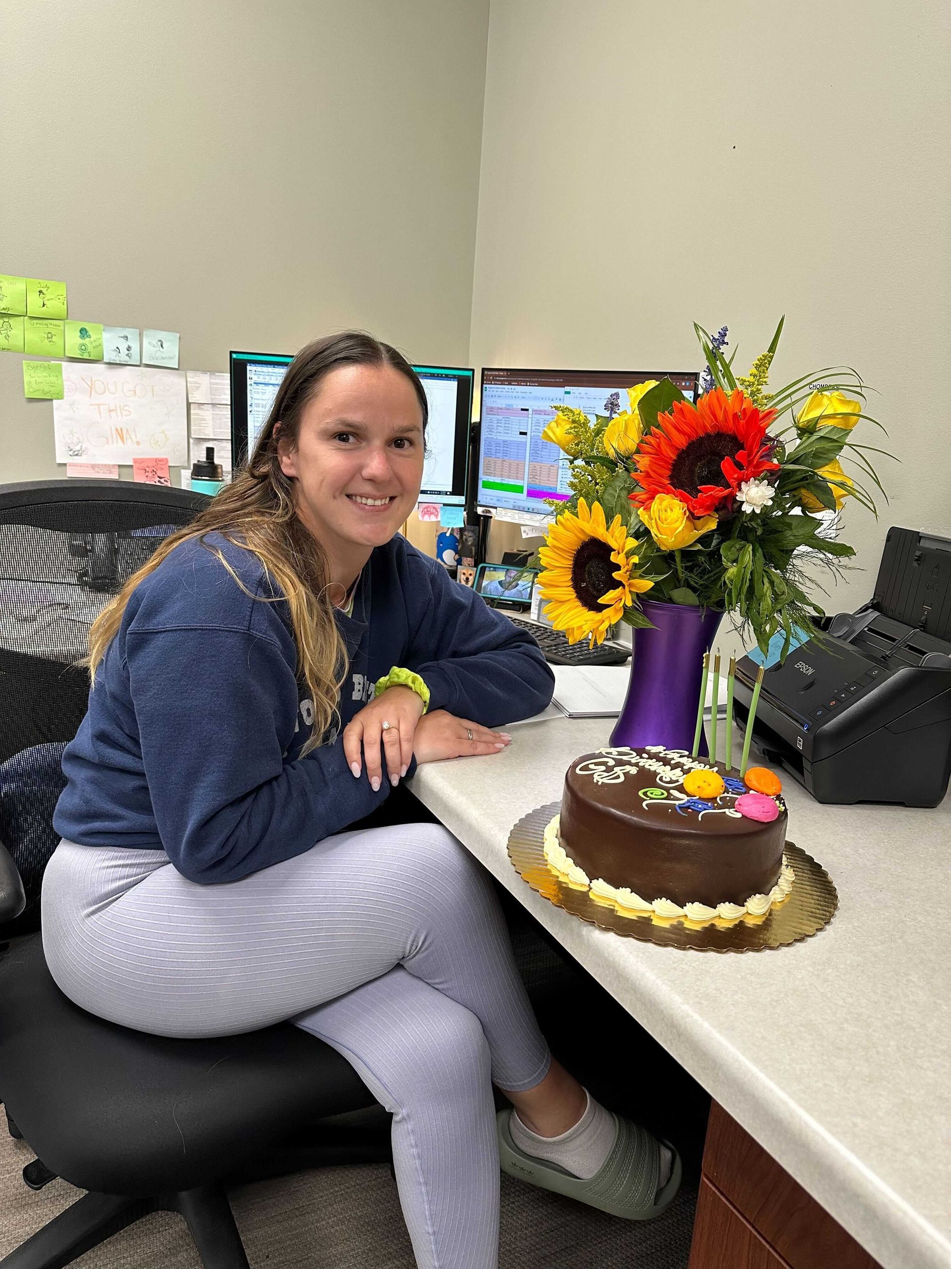 a woman is sitting at a desk with a cake and flowers on it