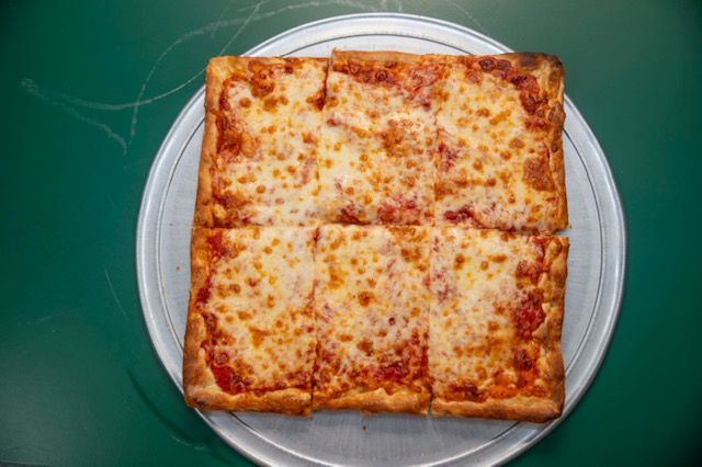 Square cheese pizza on a silver tray, cut into six slices, on a green surface.