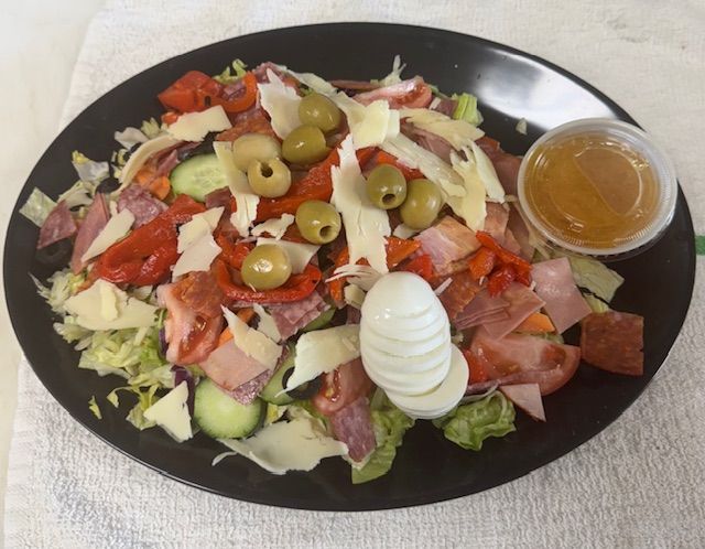 Salad with lettuce, olives, cheese, meat, cucumber, egg, and dressing on a black plate.