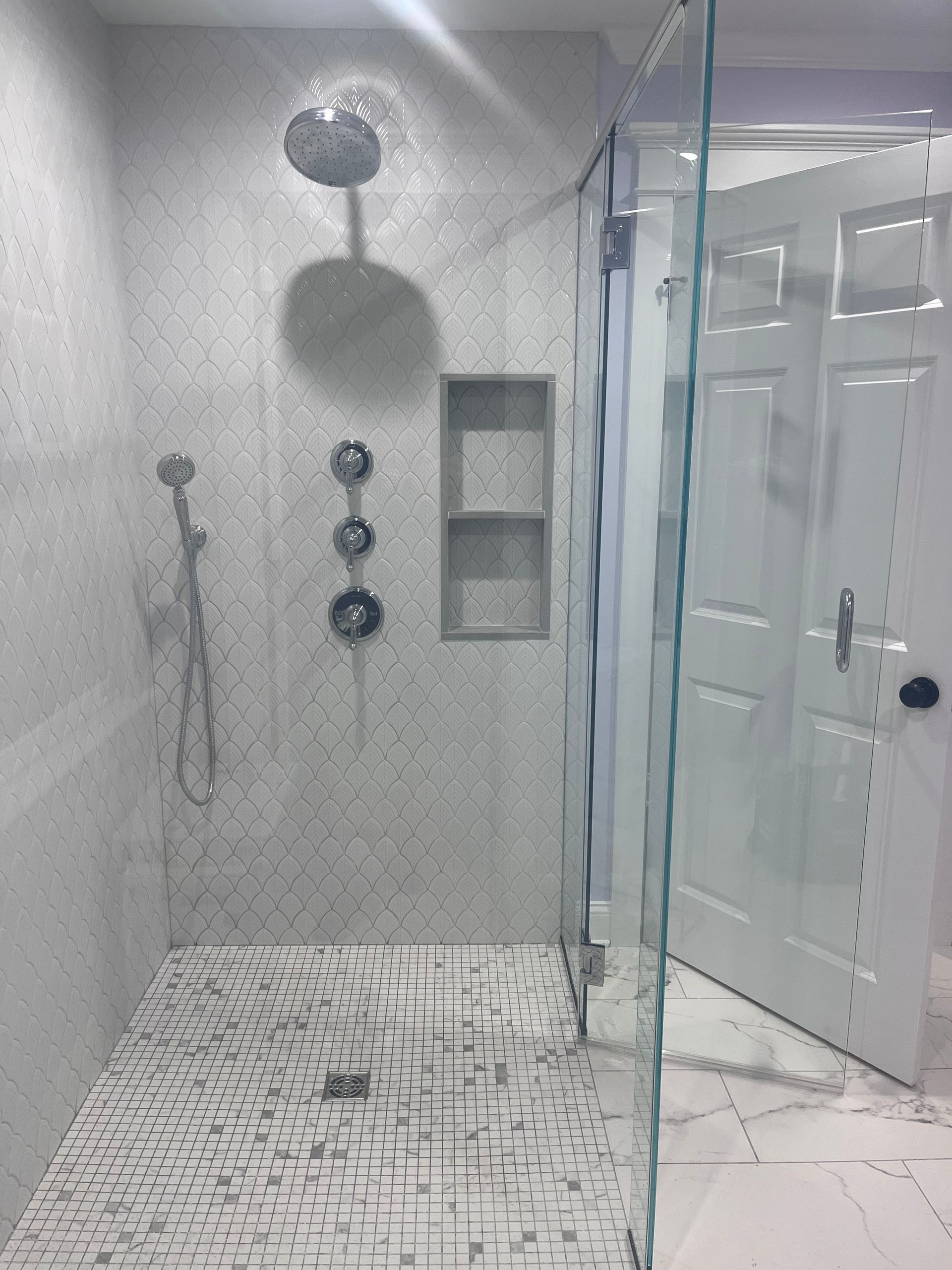 A shower stall with a mosaic-tiled wall, rain shower head, handheld sprayer, three control valves, and recessed shelving.