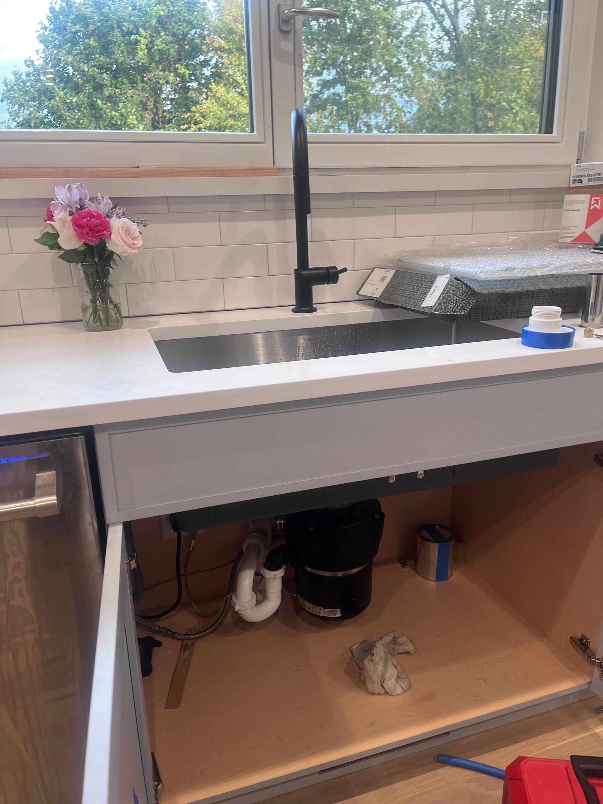 Kitchen sink with a black faucet, white countertop, and tiled backsplash, with the cabinet underneath open.