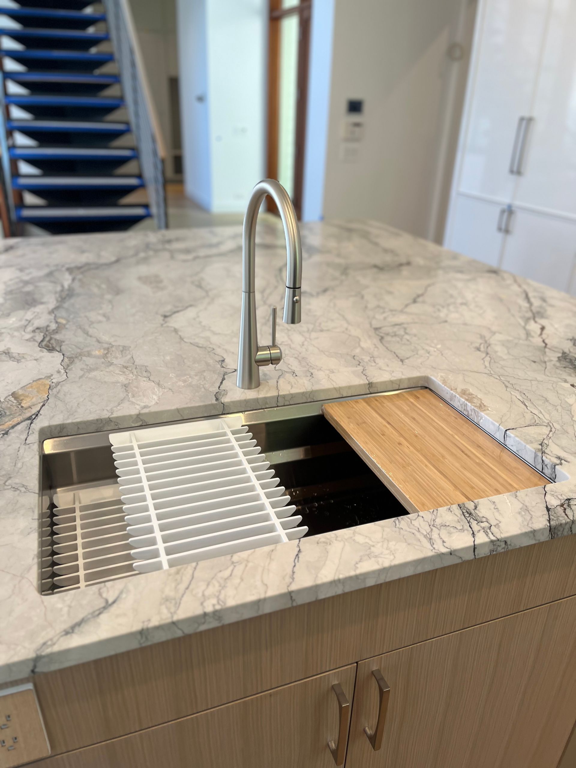 A modern kitchen island features a marble countertop, an undermount sink, a tall faucet, a rolling rack, and a wood board.