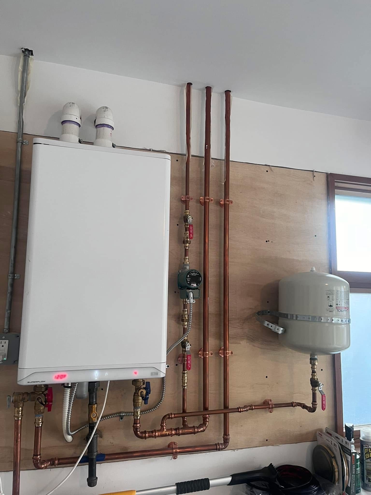 A wall-mounted boiler with exposed copper pipes and a grey expansion tank installed on a plywood panel indoors.