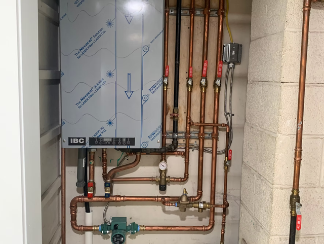 Wall-mounted gas boiler with copper pipes, valves, and gauges in a utility room.