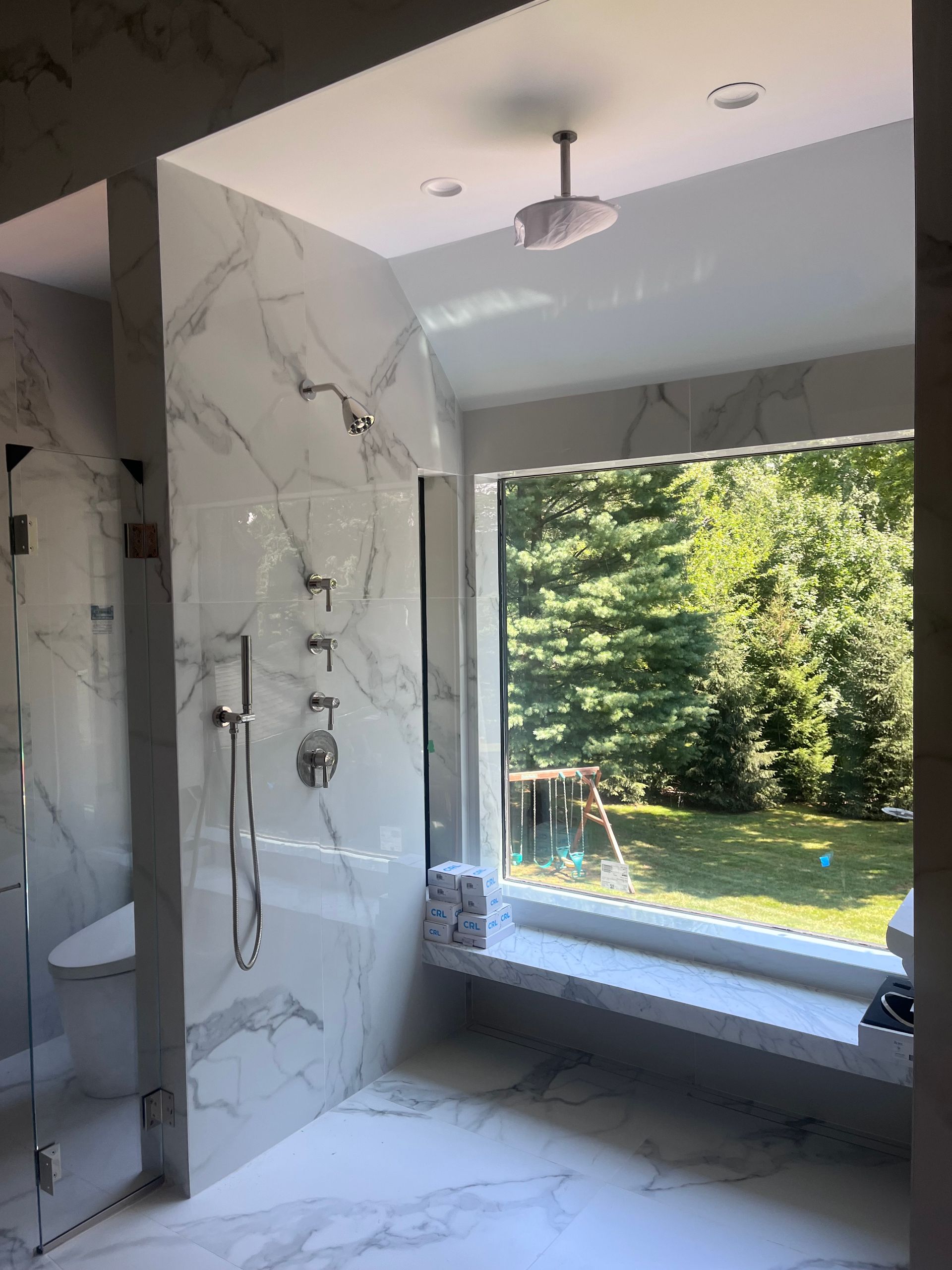 Luxurious marble-tiled shower with a large window overlooking a backyard with trees.