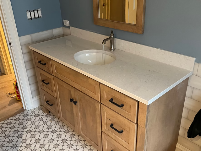 Bathroom vanity with light wood cabinets, light countertop, round sink, and patterned floor tile.