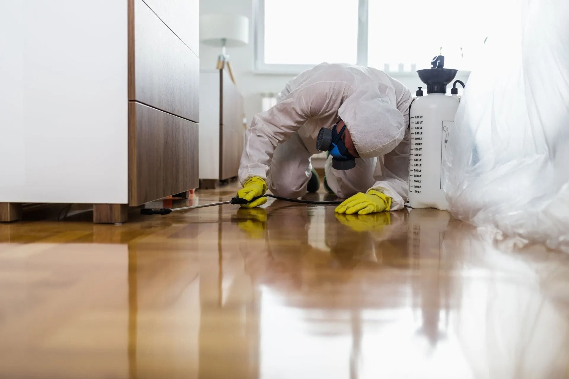 Person in hazmat suit kneeling, spraying a floor. Yellow gloves, a white sprayer, interior setting.