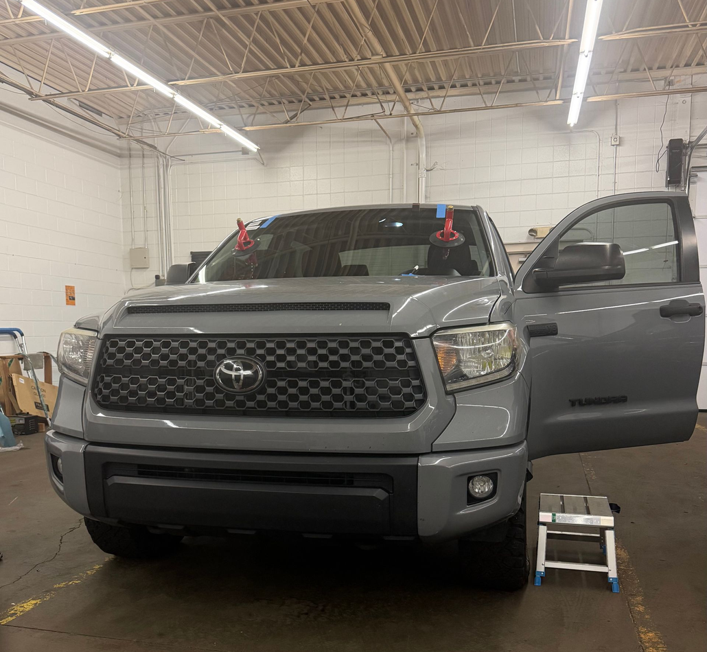 A technician in a blue shirt and black gloves uses green suction tools to install a windshield on a car in a workshop.