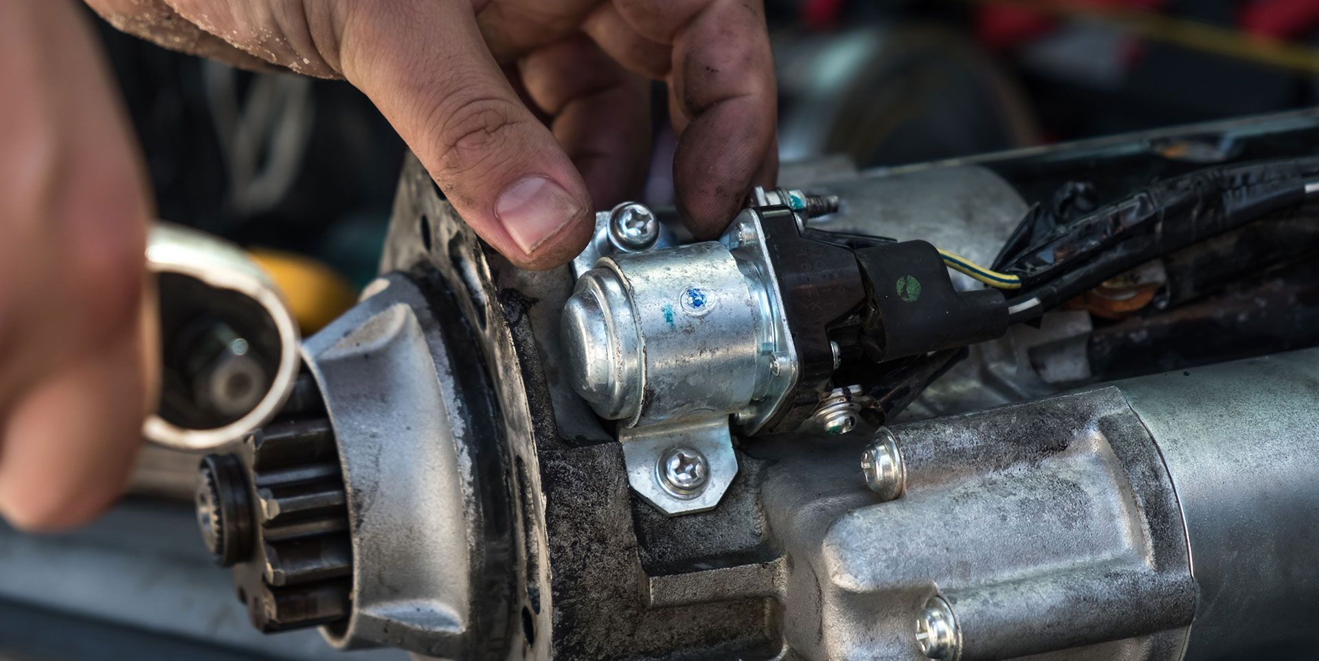 A mechanic's hands use a tool to work on a metal automotive starter motor.