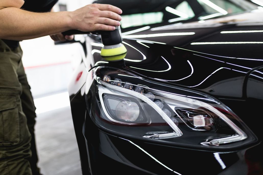 A person uses a motorized yellow polishing tool on the glossy black hood of a car near the headlight.
