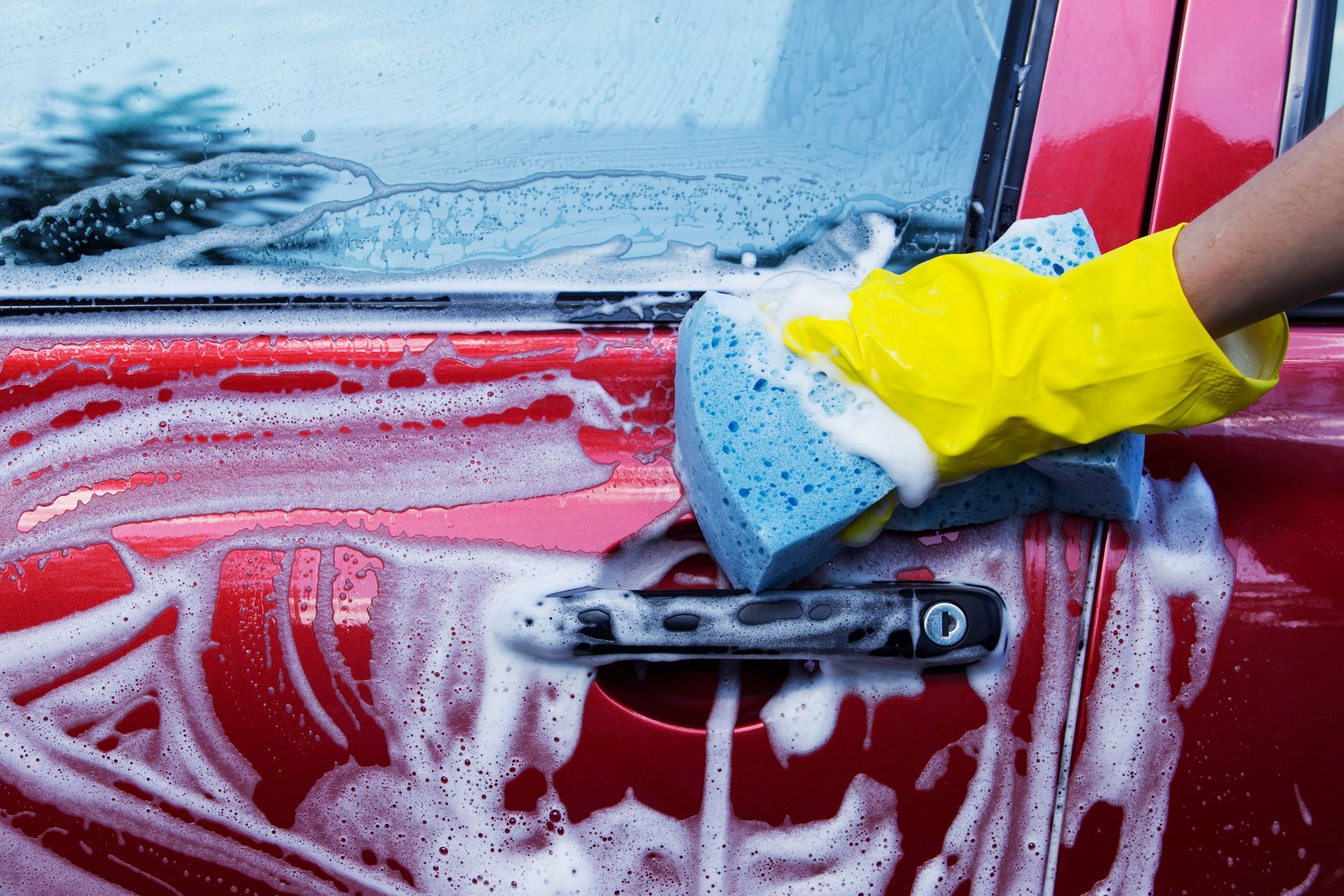 A hand in a yellow rubber glove uses a blue sponge to wash a soapy, red car door.