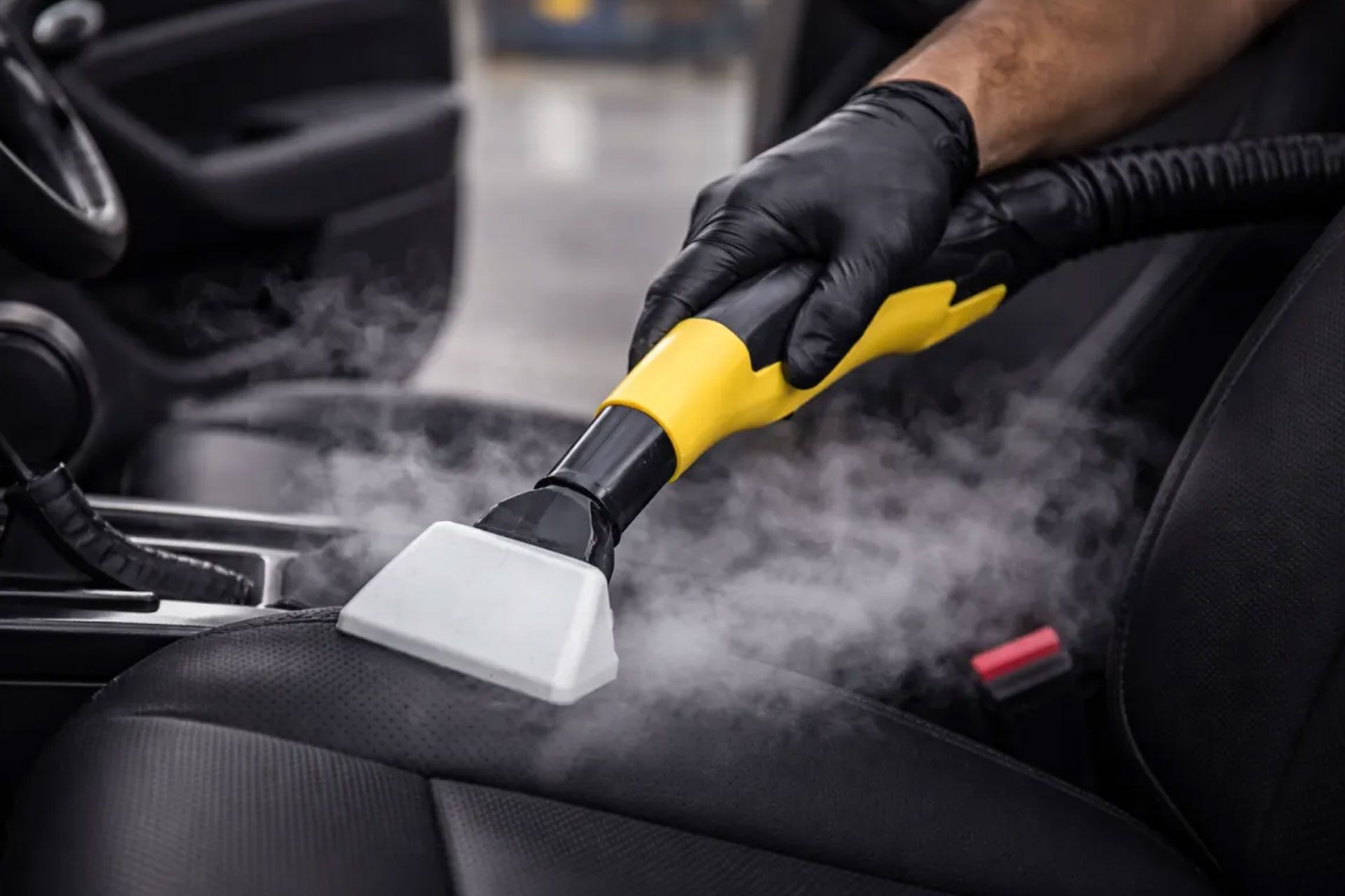A gloved hand uses a yellow and black handheld steam cleaner to sanitize a car seat.