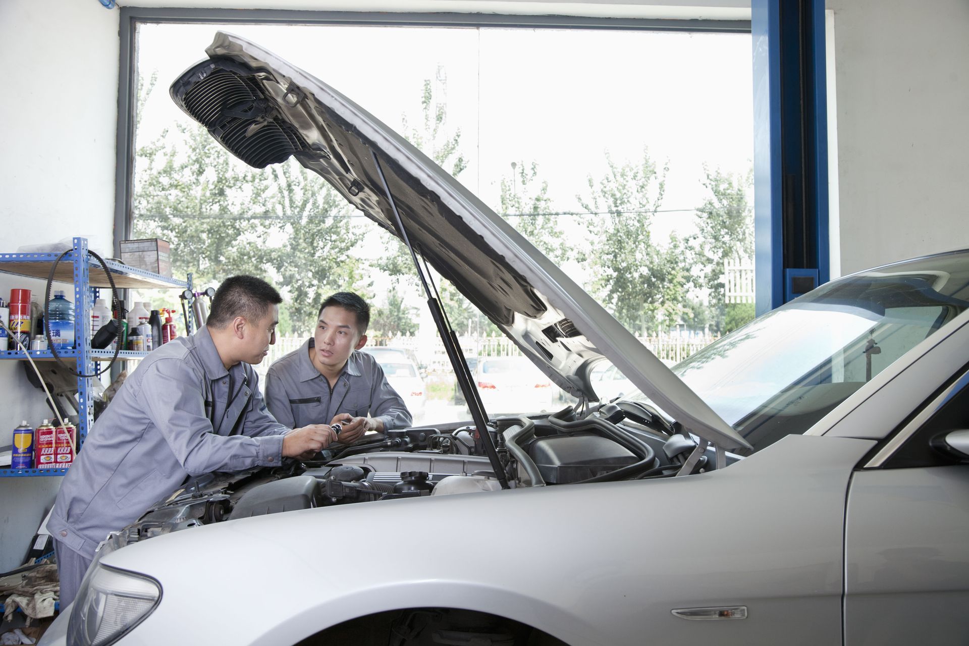 Two mechanics in uniforms examine the open hood of a silver car inside an auto repair shop.
