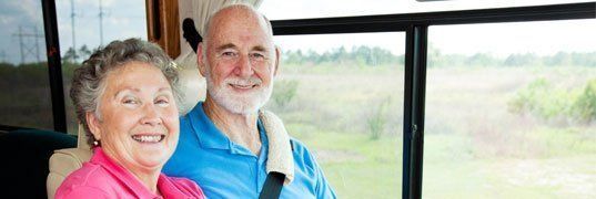 Smiling elderly couple inside a vehicle, near a large window. The man wears blue, woman pink.