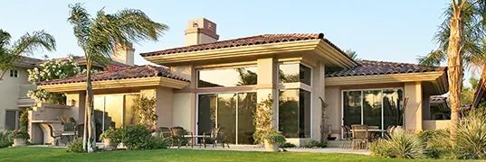 A beige single-story house with large windows, a red-tiled roof, and green grass. Palm trees are visible.