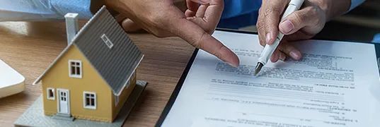 A person points to a document while signing a contract, a model house nearby on a wooden desk.