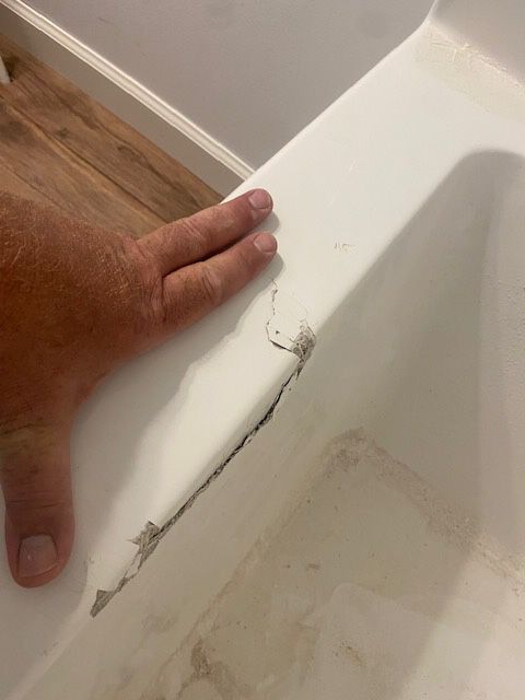 A hand touches the cracked edge of a white bathtub, revealing a dark fissure and damaged surface.