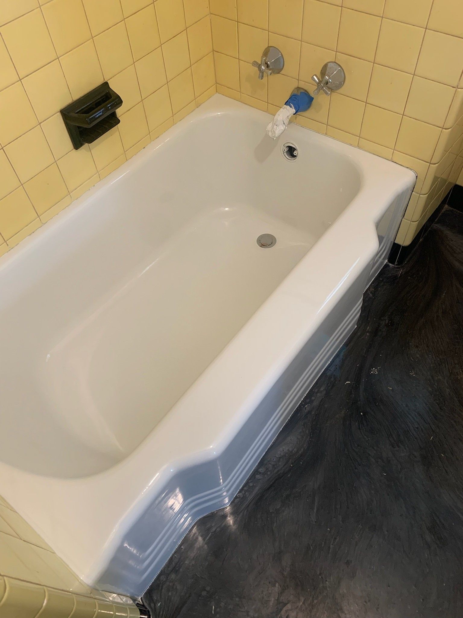 Bathtub with a white interior and light blue exterior against yellow tiled walls and a dark floor. A faucet is located at the top.