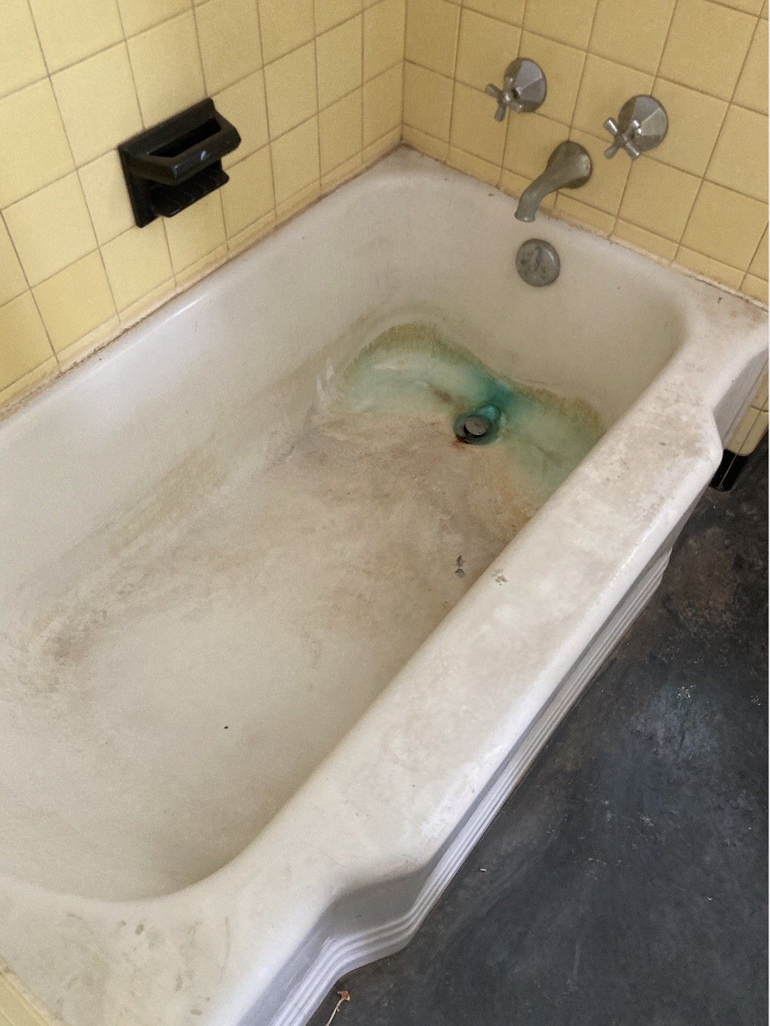 A dirty bathtub with light green water in a yellow-tiled bathroom. Soap dish is visible on the wall.