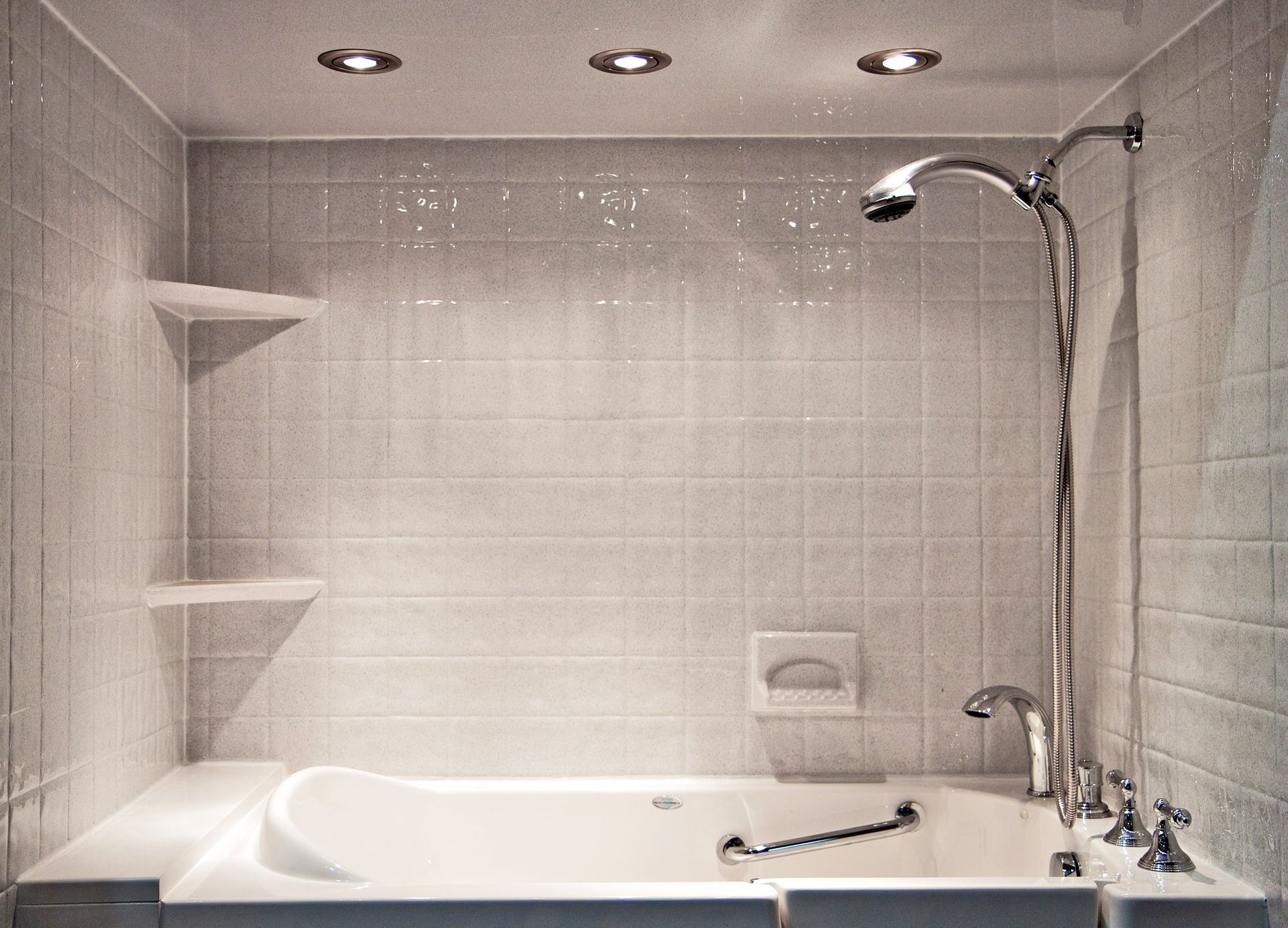 Bathroom with a white bathtub, showerhead, and tiled walls. Built-in shelves are on the left, and recessed ceiling lights illuminate the space.
