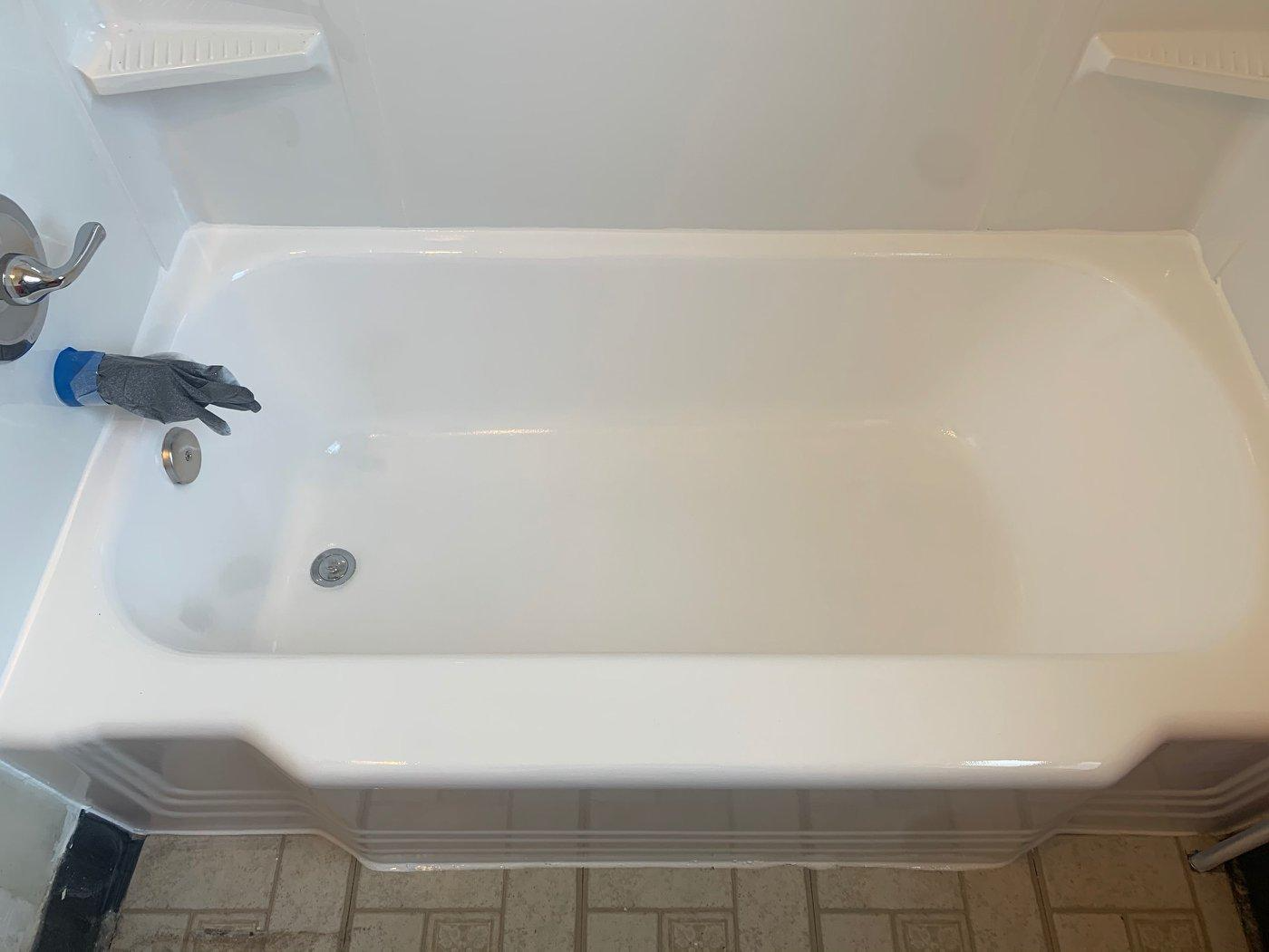 A white bathtub with a faucet, surrounded by white walls and a tiled floor. A hand in a glove is near the faucet.