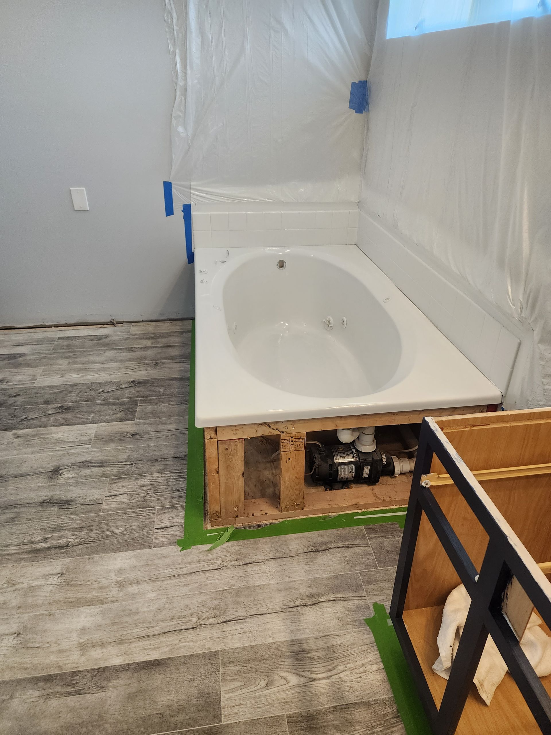 Bathroom renovation in progress: a white jacuzzi tub sits in a wooden frame. The floor is gray laminate. Plastic sheeting covers the walls.