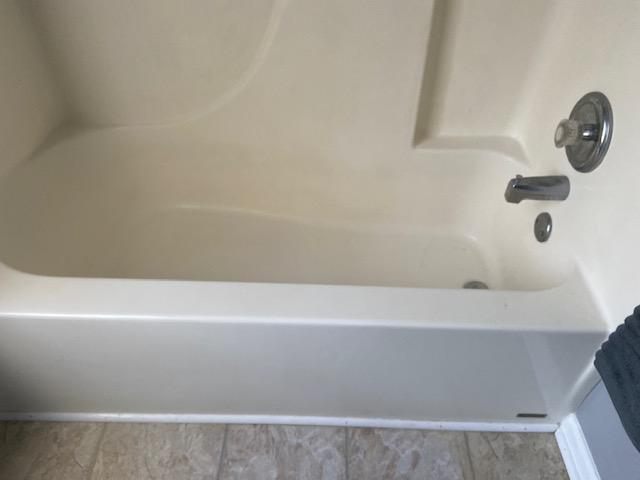 An ivory-colored bathtub with a curved interior and silver faucet. It is situated against a tiled wall, and next to a section of gray flooring.