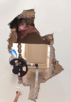 Hole in a wall revealing plumbing fixtures, including pipes and a black valve. Beige wall and a glimpse of a room behind the hole.