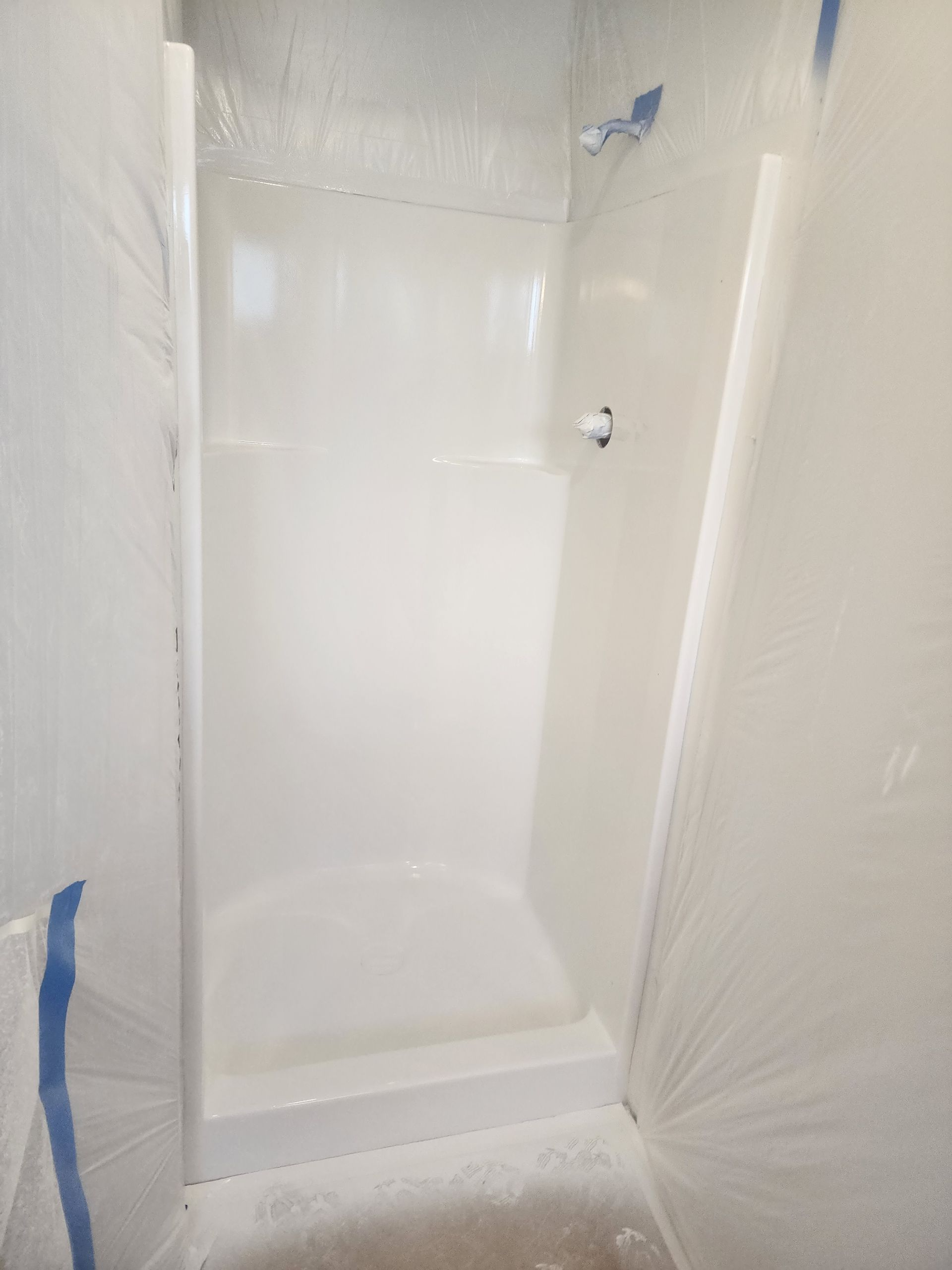 A white shower stall enclosed by plastic sheeting secured with blue tape. A shower head and faucet are visible.