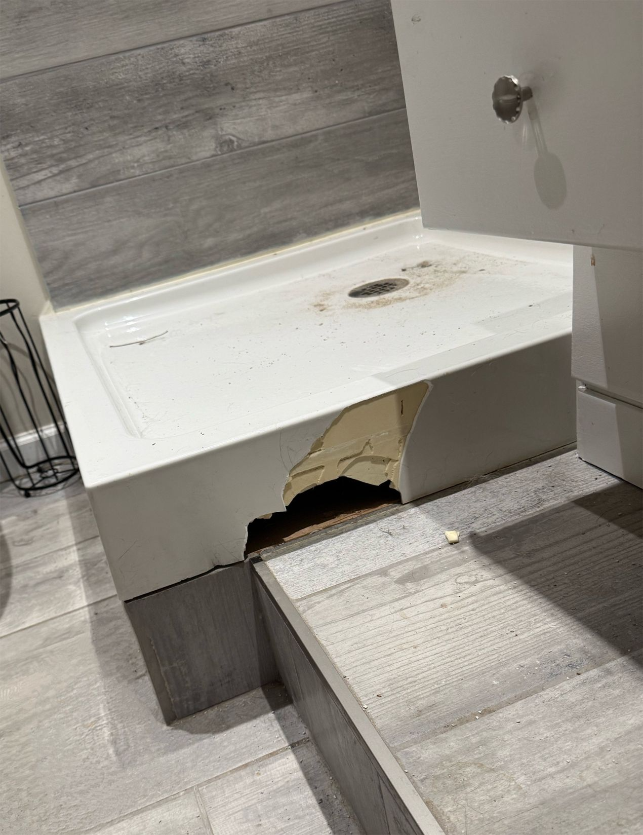 Damaged white shower base with a large hole in the side, next to a step and toilet. Gray tile surrounds.