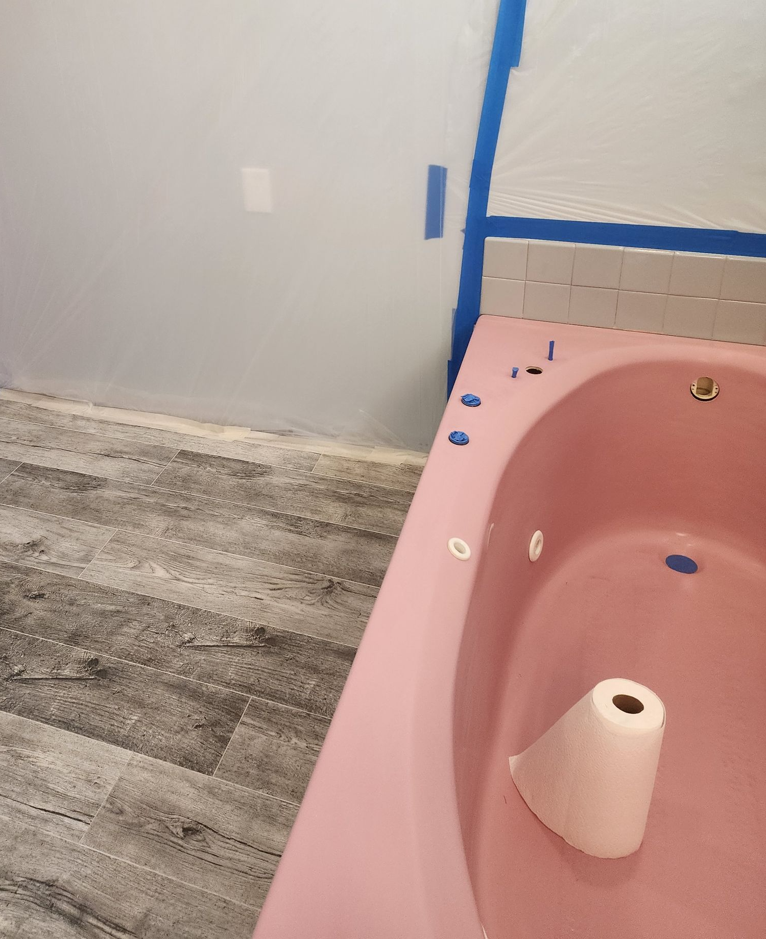A pink jacuzzi bathtub with grey tile trim and gray wood-look flooring. Plastic sheeting is taped to the surrounding wall.