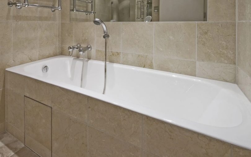 A white bathtub built into a beige tiled wall. The shower head hangs above.
