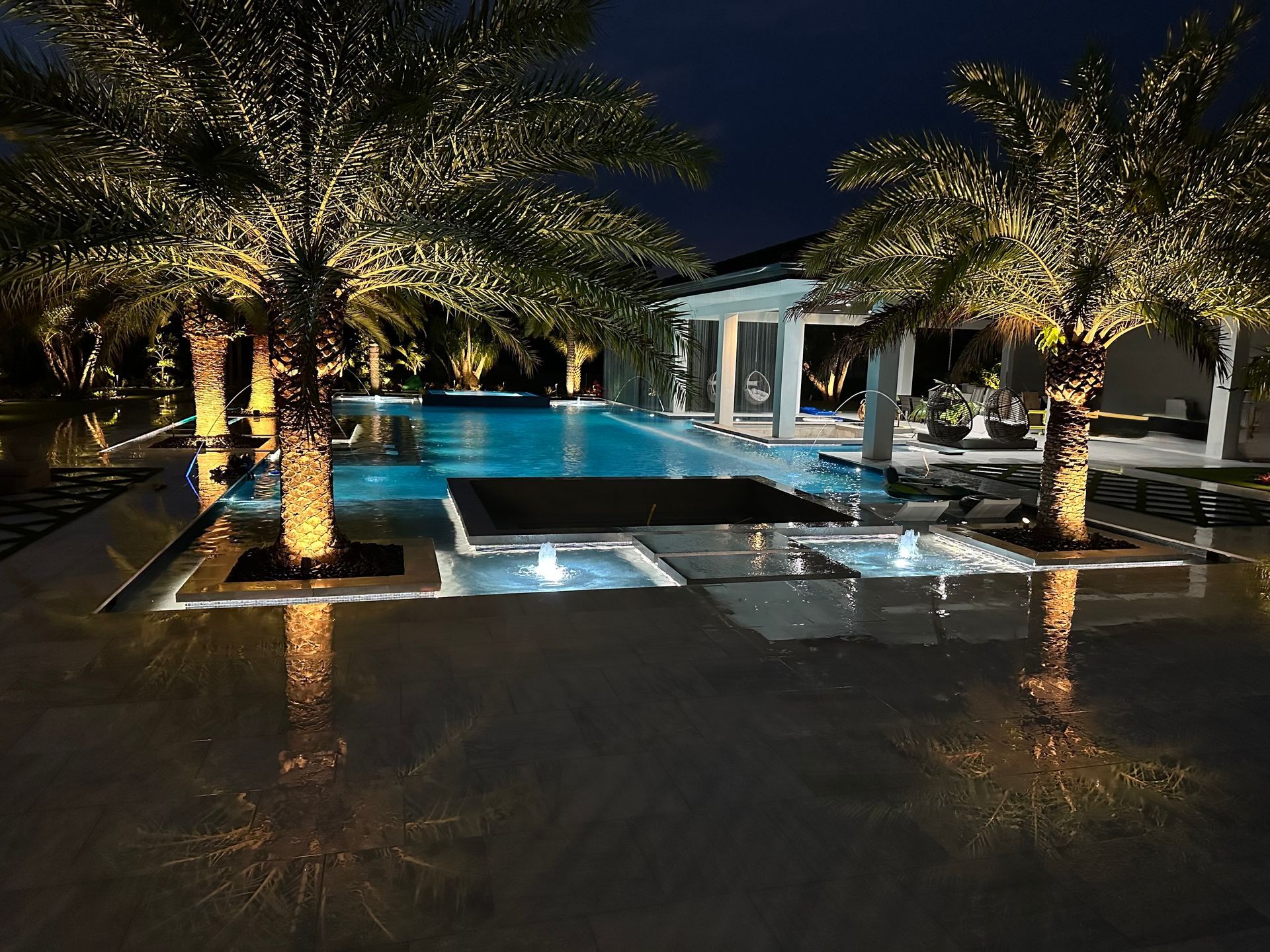 A large swimming pool is lit up at night surrounded by palm trees