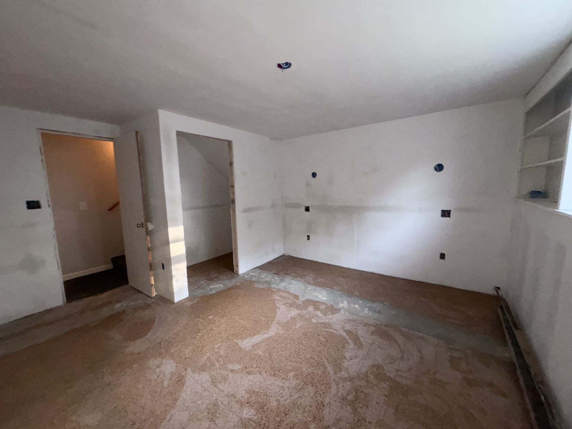 Interior of a room under construction, with white walls, exposed wiring, and stained floor.