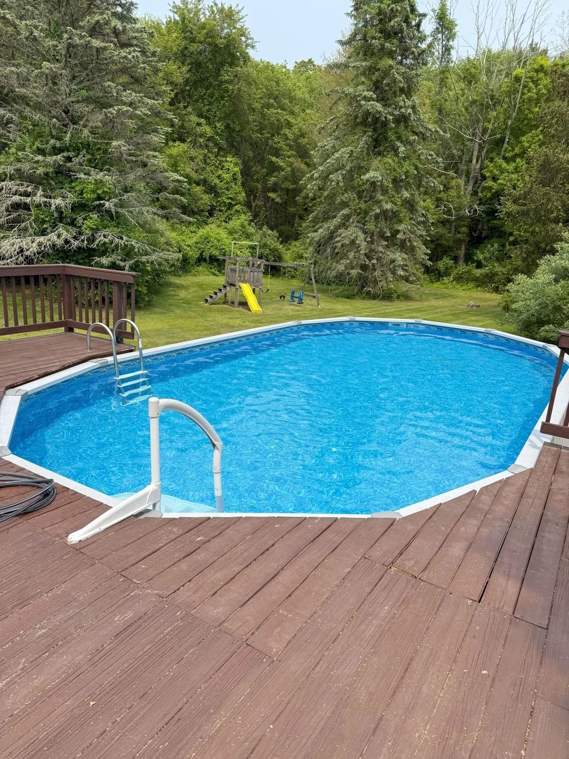 An above-ground pool on a wooden deck, surrounded by trees and a green lawn.