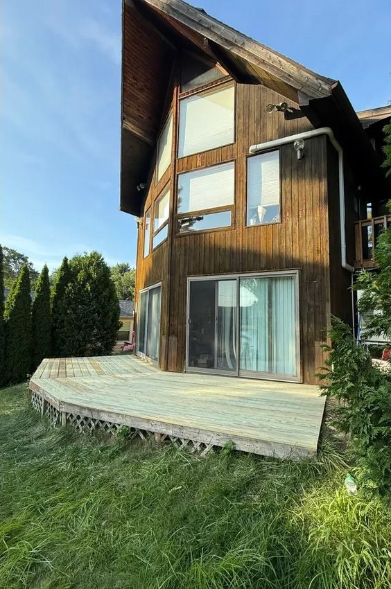 Wooden house with large windows, sliding glass doors, and a deck on a grassy lawn.