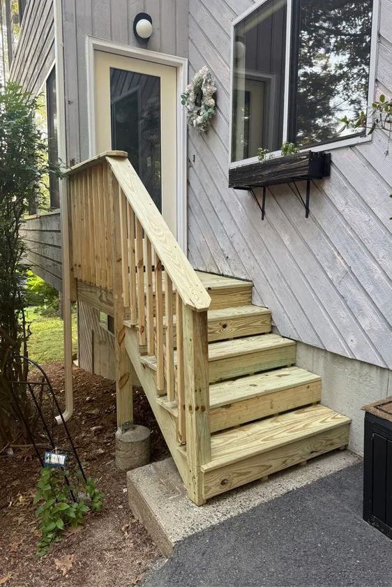 Wooden stairs leading up to a white door, with a railing. Gray siding and a window with a planter.